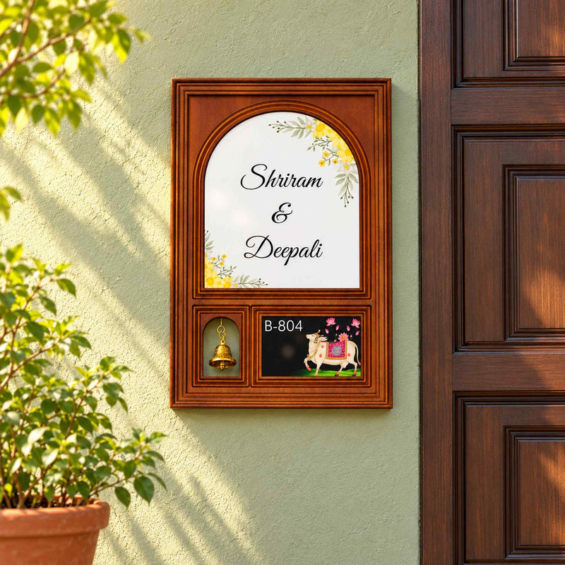 Shriram and Deepali floral wooden nameplate with hanging bell and B-804 cow artwork on a sage-green entrance wall.
